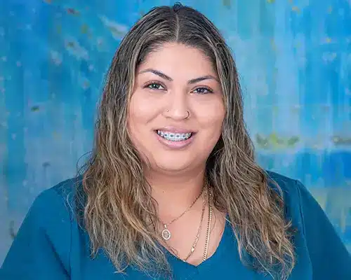 Professional headshot of smiling woman with long brown hair against blue background for orthodontics practice.