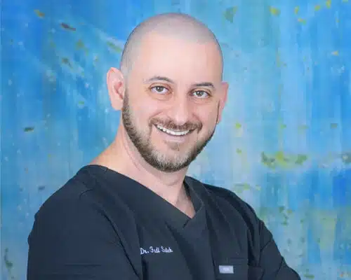 Professional headshot of smiling man in black scrubs against blue medical office background.
