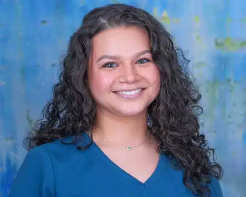 Young woman smiling at camera wearing blue shirt against blue background for orthodontics practice.
