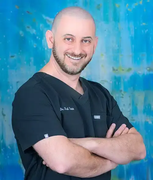 Smiling male orthodontist in black scrubs with arms crossed against blue background.