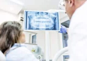 Photo of health dental professional in blue scrubs examining dental x-ray on computer screen, isolated dentist office. People, medicine, technology and health care concept - dentists looking to x-ray scan on monitor at dental clinic. Rear view of male dentist explaining the details of x-ray to a beautiful female patient