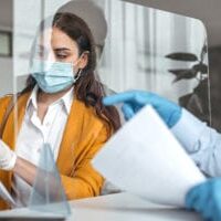 Female orthodontist in yellow jacket wearing mask examines patient documents at desk with protective barrier.